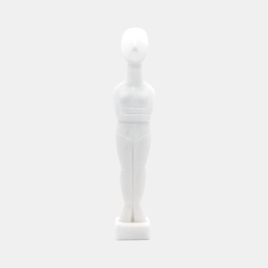 The image shows a stylised white statue or figurine made from White Naxos Marble. It is a simplified representation of a human figure with an abstract and geometric design. The figure has no facial features and stands upright on a small base. The aesthetic is minimalist, focusing on form and shape rather than detail.