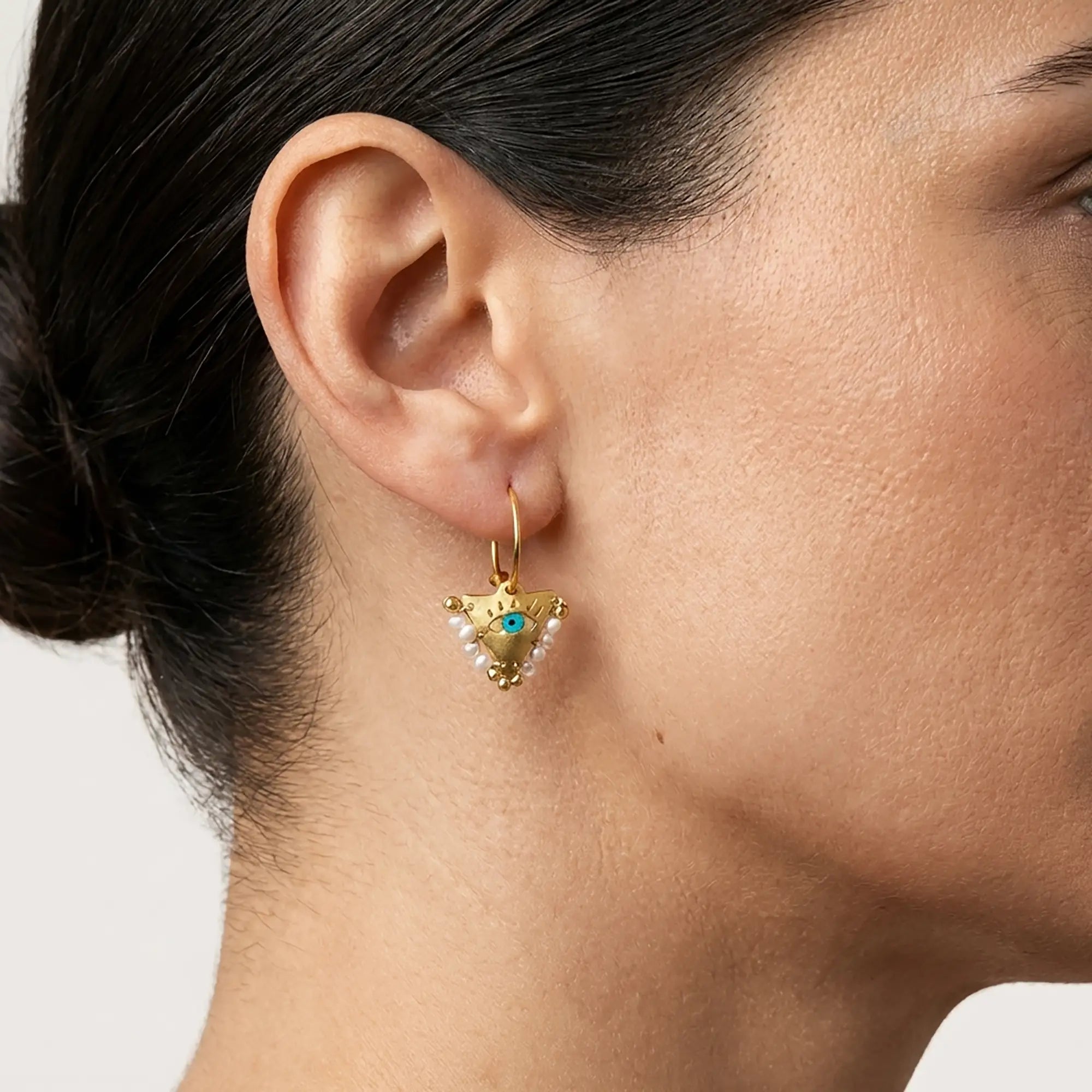 Woman wearing small white pearl Fylachto earrings in gold plated sterling silver with evil eye detail