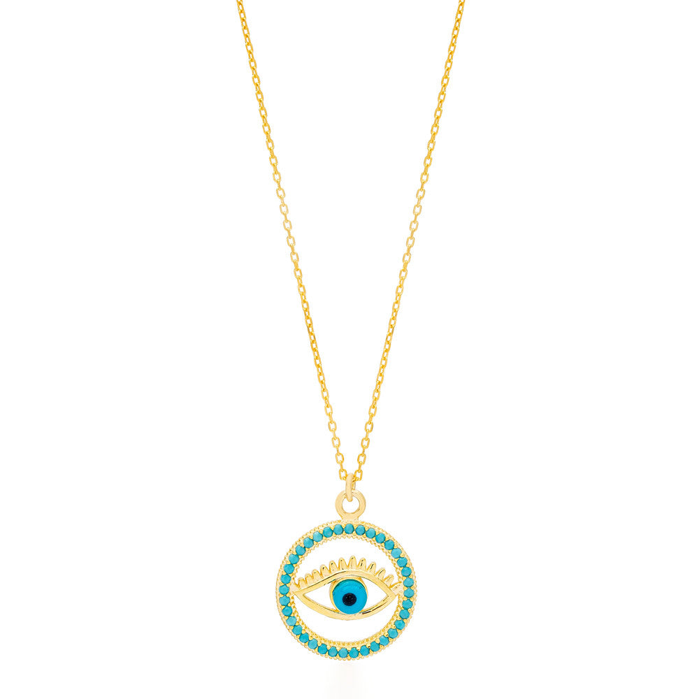 The image shows a gold-colored necklace with a pendant that features the design of an eye. The eye is centered within a circular frame, and the frame is embellished with what appears to be turquoise-colored stones or crystals. The eye itself has a blue iris, which stands out against the gold and turquoise colors. This type of design is often associated with the 