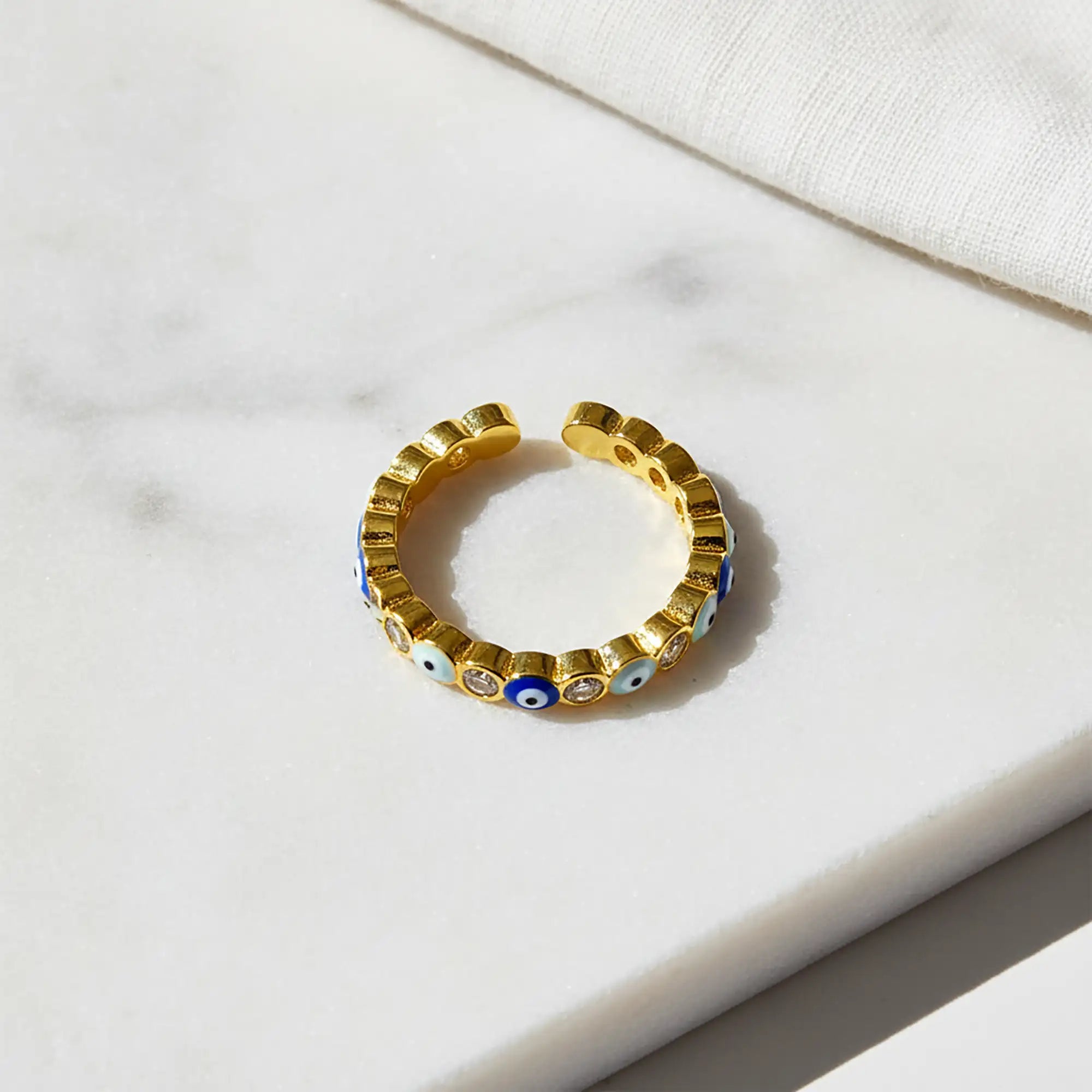 Adjustable gold evil eye ring with alternating blue enamel eye motifs and sparkling clear crystals on a white marble surface