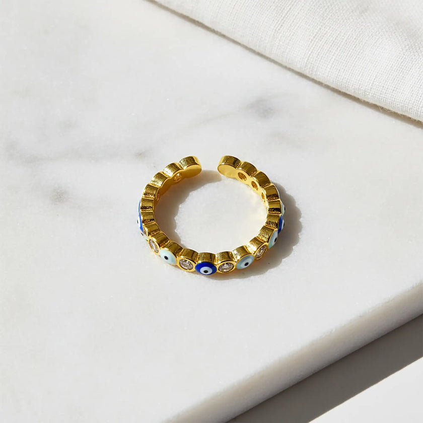 Adjustable gold evil eye ring with alternating blue enamel eye motifs and sparkling clear crystals on a white marble surface
