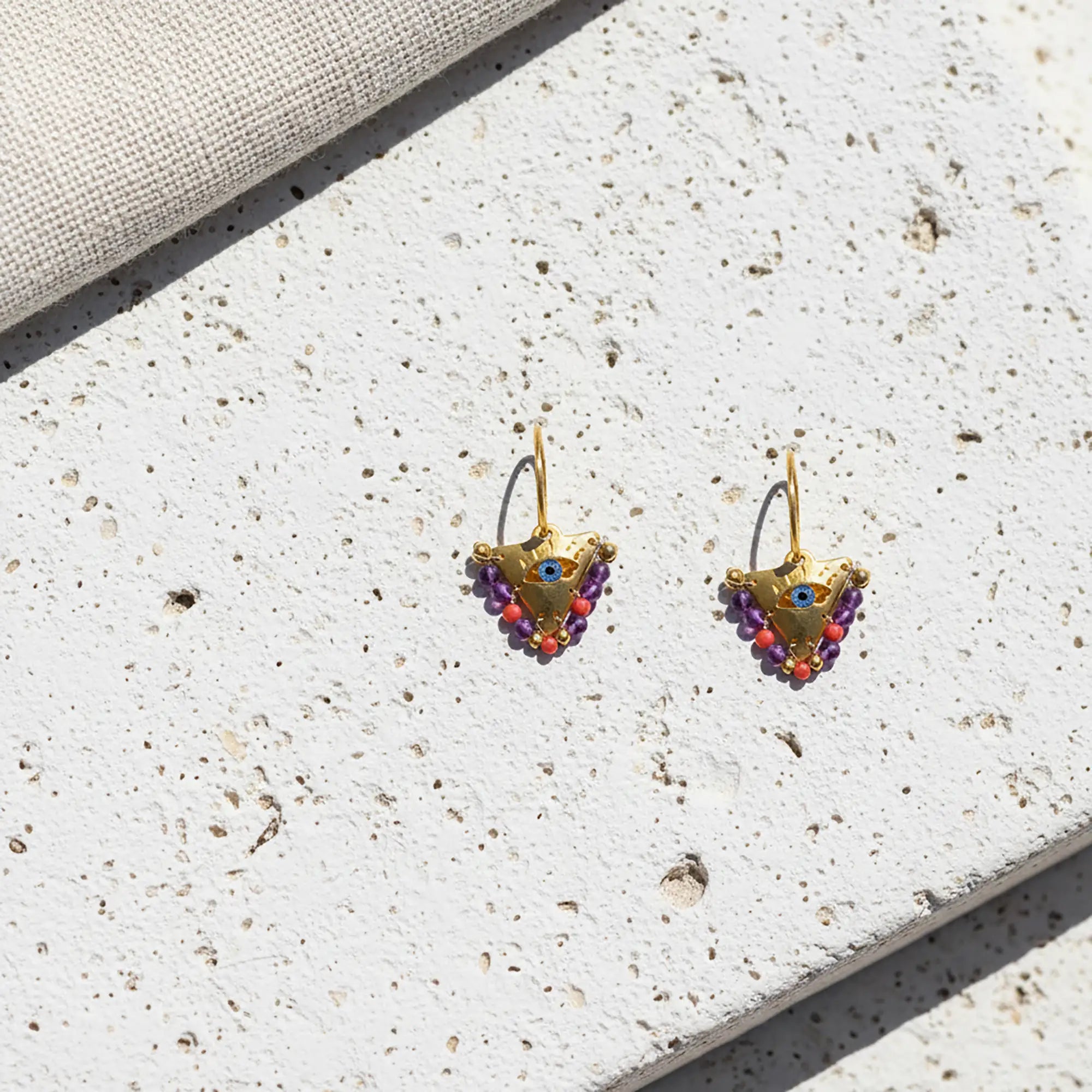 Pair of colorful small fylachto earrings with handpainted blue eye and purple coral bead details on light stone surface