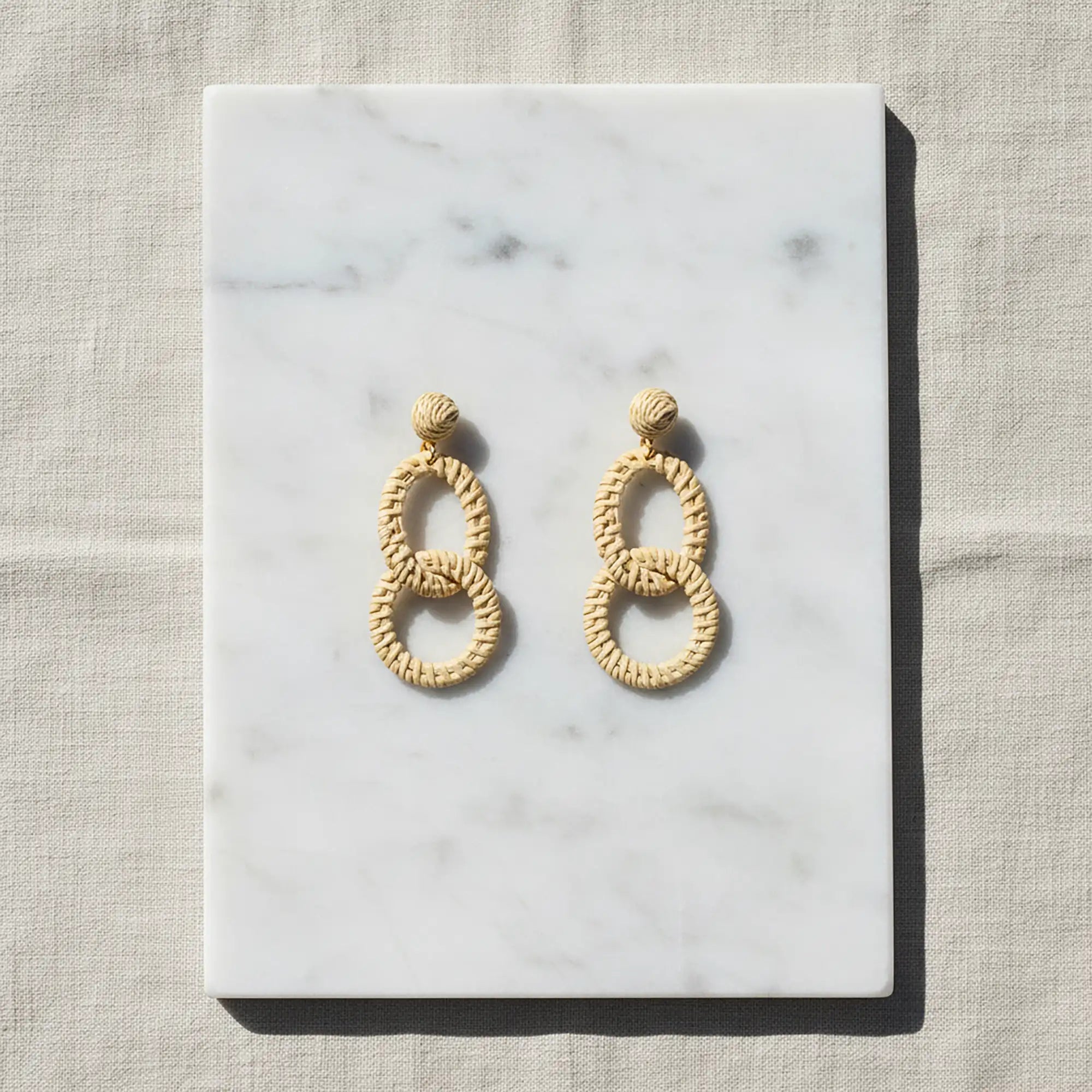 Natural woven double oval earrings displayed on white marble with soft neutral fabric