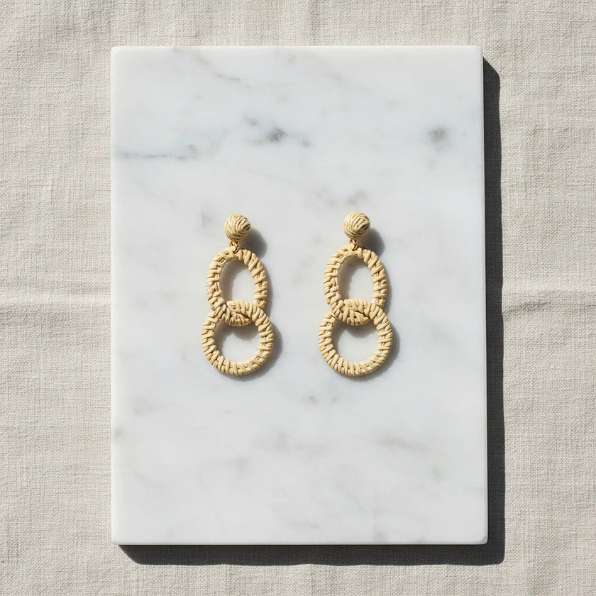 Natural woven double oval earrings displayed on white marble with soft neutral fabric